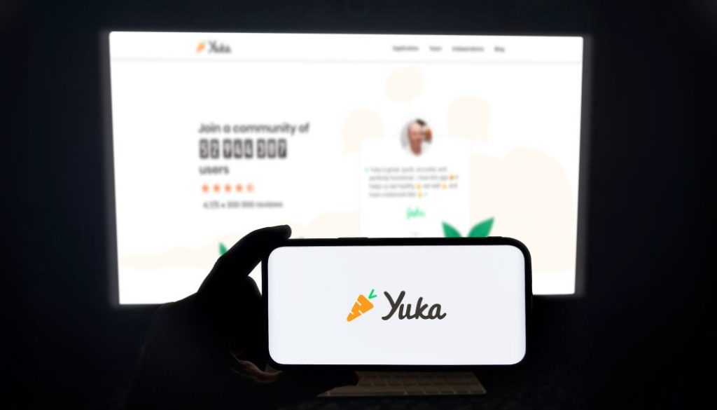 Yuka app running on a modern mobile phone. The smartphone is being held in landscape mode, in the darkness. A PC monitor with the Yuka website in the background.