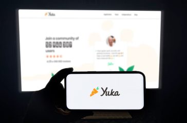 Yuka app running on a modern mobile phone. The smartphone is being held in landscape mode, in the darkness. A PC monitor with the Yuka website in the background.