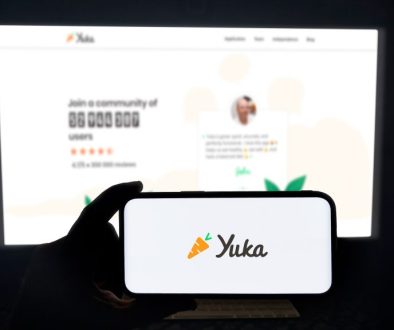 Yuka app running on a modern mobile phone. The smartphone is being held in landscape mode, in the darkness. A PC monitor with the Yuka website in the background.