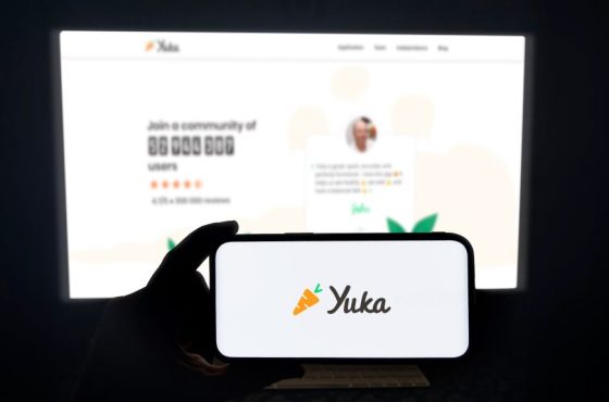 Yuka app running on a modern mobile phone. The smartphone is being held in landscape mode, in the darkness. A PC monitor with the Yuka website in the background.