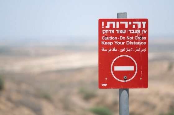 A bright red warning sign outside of the Gaza Strip, near the separation fence around the territory. The sign is trilingual, in Hebrew, Arabic, and English.