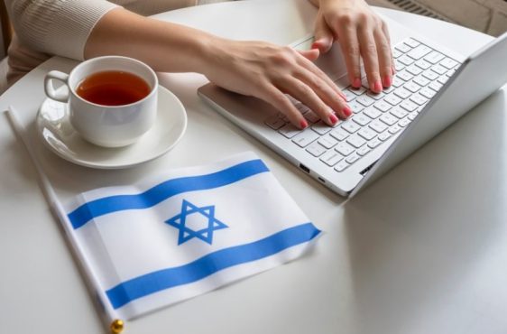 Lonely woman freelancer with flag of Israel enjoying having breakfast with cup of coffee working on laptop sitting near window in cafe at morning.
